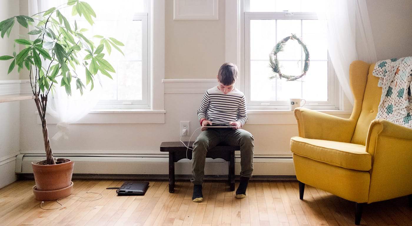 A boy sitting in his living room on a wooden bench looking at his tablet device