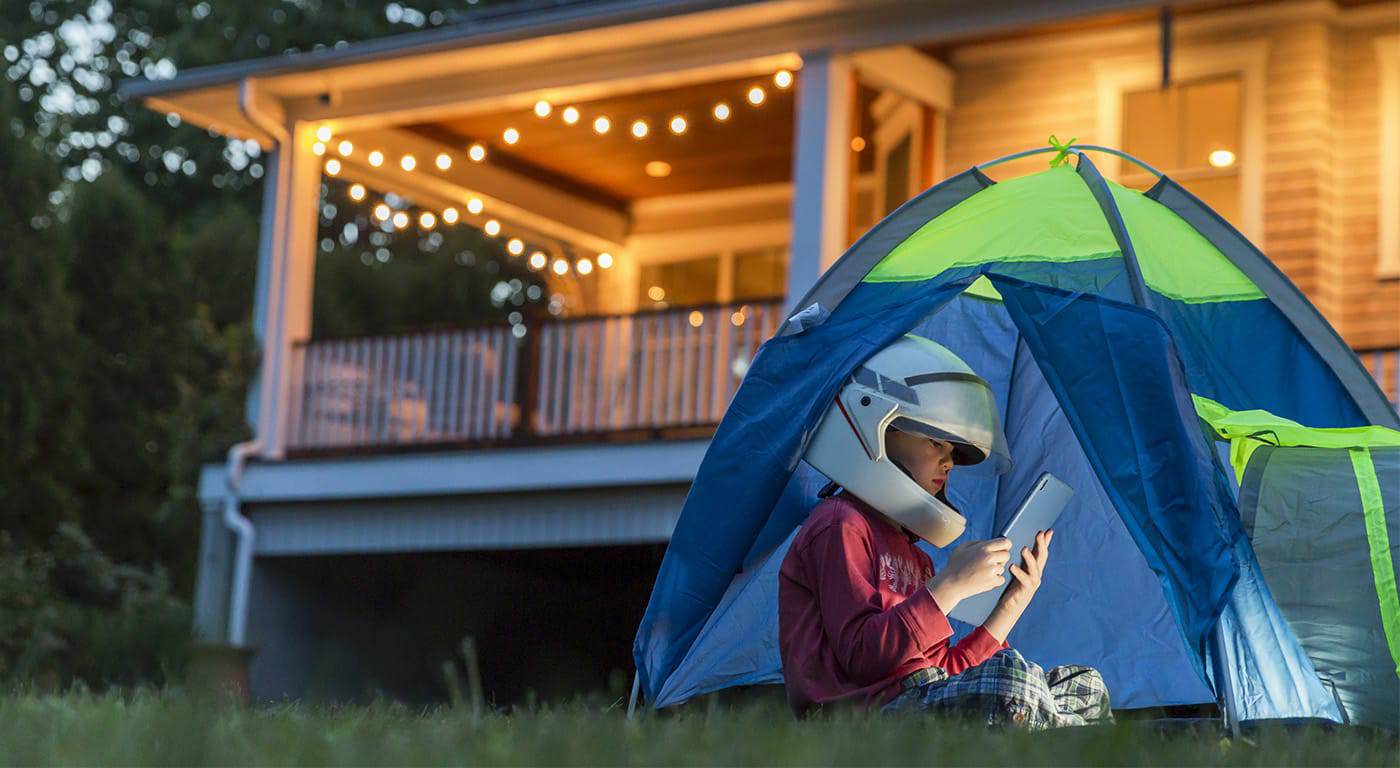 Little boy sitting outside in a blue tent with an astronaut helmet on looking at his iPad