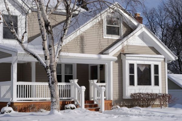 A light gray house with white trim and a large front porch is covered in snow. A bare tree stands in front, and icicles hang from the roof, reflecting the bright sunlight of a clear winter day.